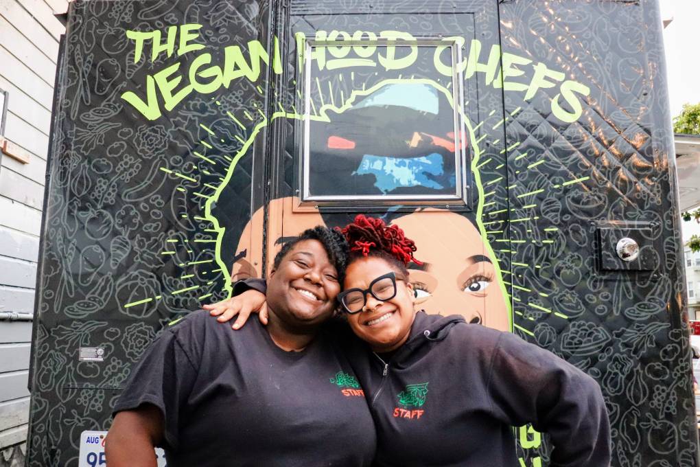 two women, known as the Vegan Hood Chefs, hugging in front of a food truck with the logo "The Vegan Hood Chefs"