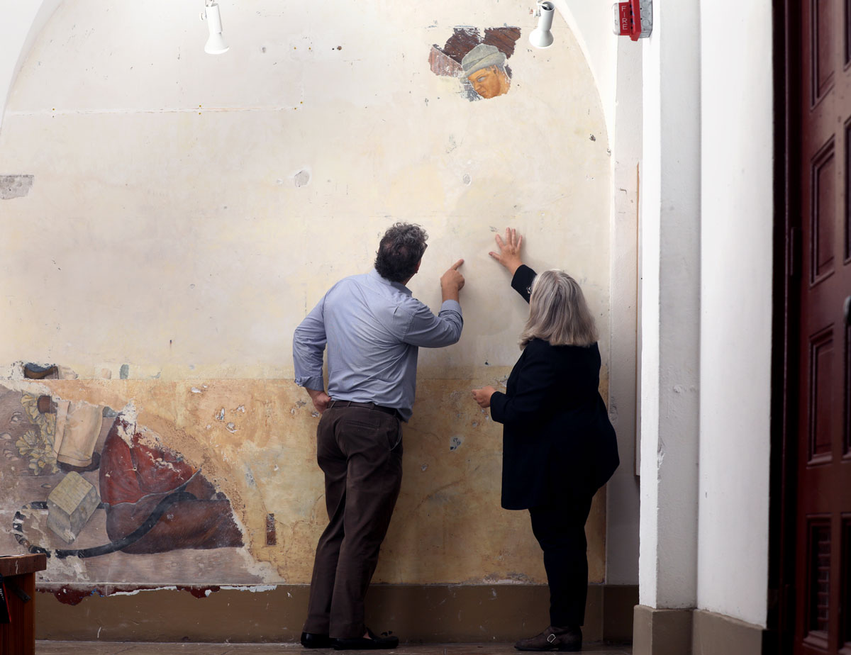 Two figures with their backs to camera point at wall with some revealed painting portions