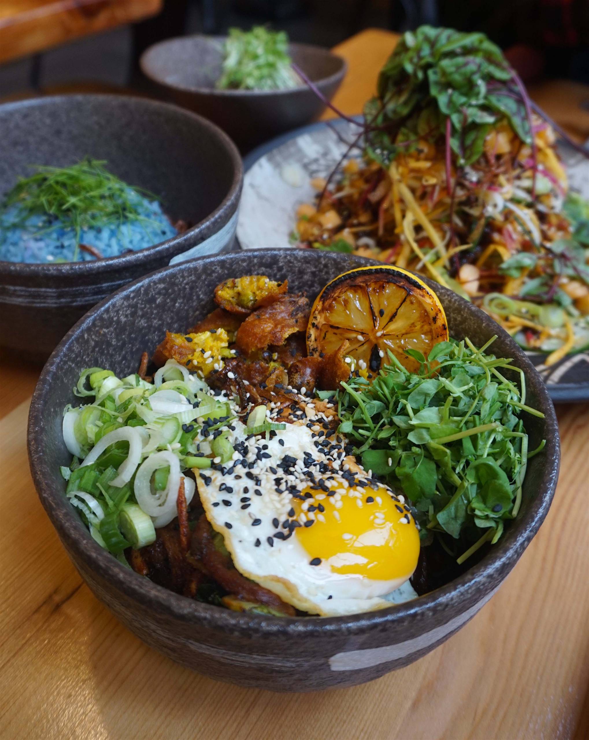 In the foreground, a bowl of turmeric noodles topped with greens, scallions, a charred lemon, and a fried egg.