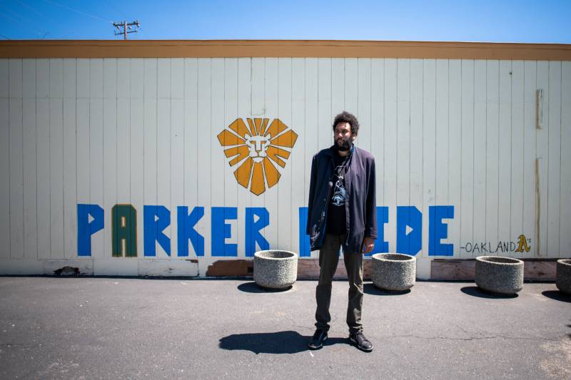 Tongo Eisen-Martin stands outside Parker Elementary on a sunny day. He is wearing a blue peacoat and green pants. The wall behind him says "Parker Pride"