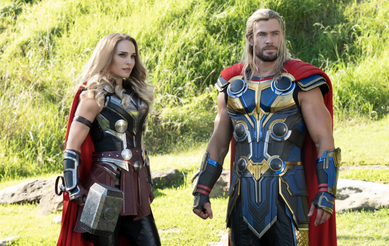 A blonde and muscular man and woman stand in a field of green grass, wearing shiny superhero armor. She clutches a giant hammer. He wears a red cape.