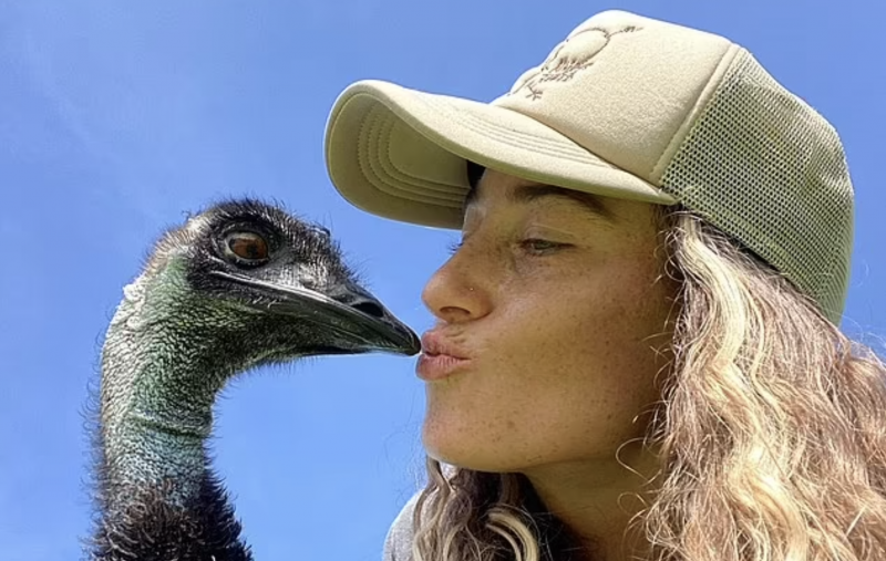A young woman with long wavy blonde hair puckers her lip against the pointed end of an emu's beak. She is wearing a beige baseball cap.