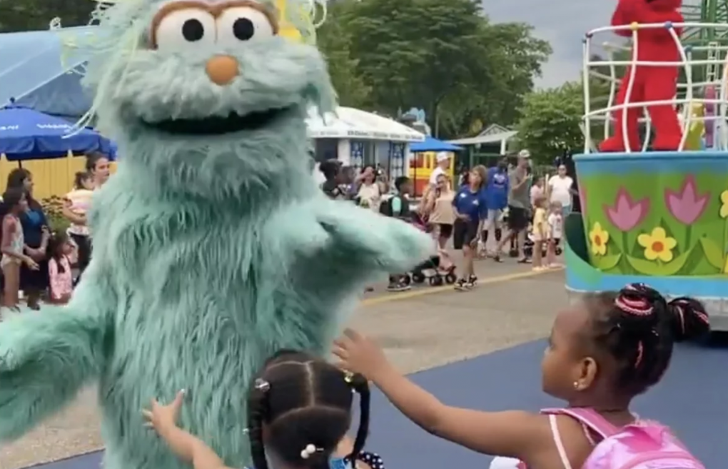 An adult wearing a green Sesame Street character costume passes by two little Black girls who are reaching out for hugs.