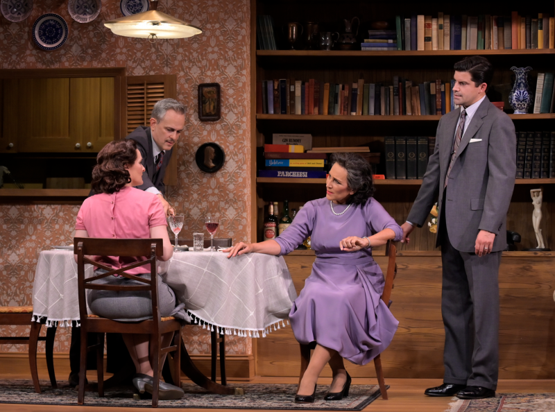 A woman in a lilac dress sits at a dining table and speaks to another seated woman, wearing grey skirt and pink shirt. To her right is a young man wearing a grey suit. To her left, an older man in a grey suit pours a drink at the table. All are wearing period clothing from the 1950s. Behind them are shelves full of books and boardgames.