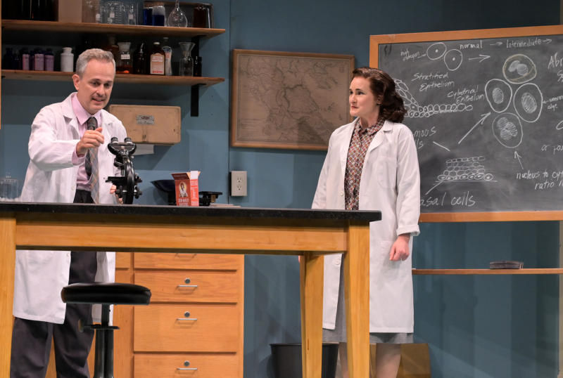 A grey haired man and a young woman, both wearing white lab coats work in a lab. In front of them is a table with a microscope on it. Behind them a black board with diagrams of cells on it and shelves full of medical bottles.