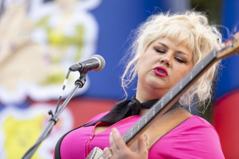 A woman with a big blond '60s-inspired hairdo looks down at her guitar, behind a mic stand. She is wearing a bright pink blouse and matching lipstick