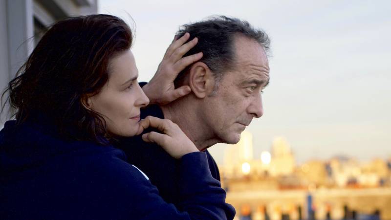 A man peers over a balcony, a city gleaming in the sun behind him. At his side a woman leans on his shoulder, affectionately caressing the back of his head.