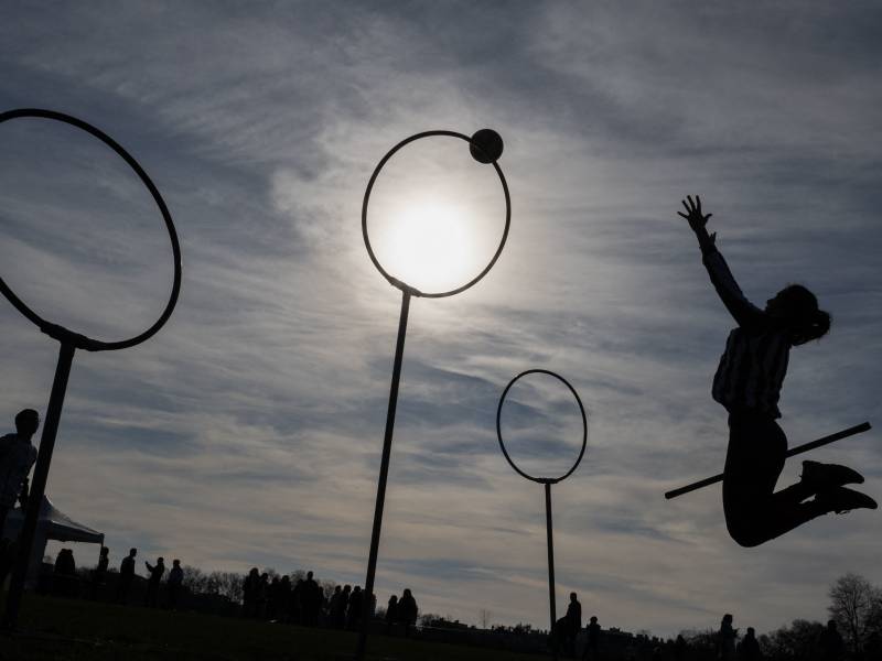 A woman, in silhouette, is seen leaping towards a round hoop on a stick. She has tossed a ball towards it. 