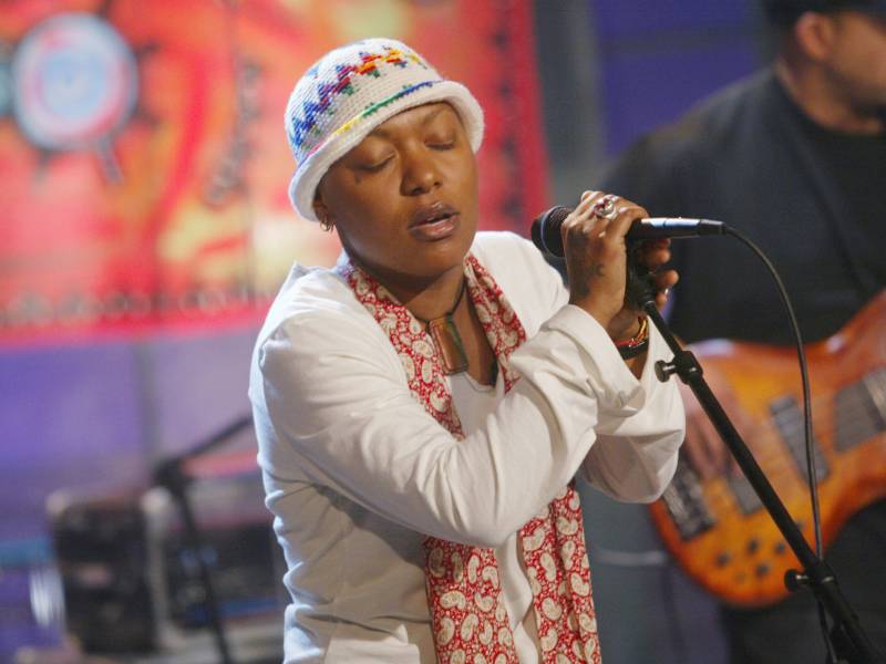 A Black woman wearing a white embroidered bucket hat, a white sweater and flowing red and white scarf takes a pause for breath, while holding a microphone attached to a stand, mid-performance.
