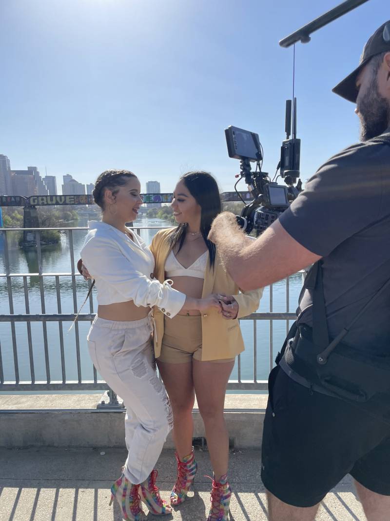 two salsa dancers perform in light clothes on a bridge against the backdrop of Austin, Texas, while a videographer films