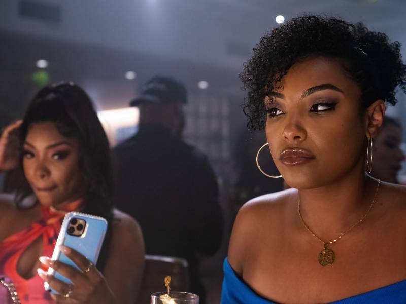 Two Black women sit in a nightclub wearing form-fitting dresses. Both have serious facial expressions. One is holding up a cell phone. 
