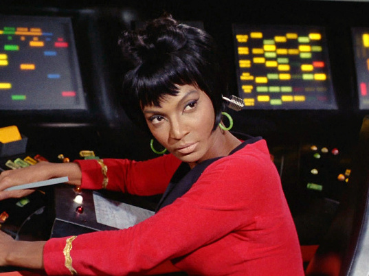 A beautiful Black woman with hair styled into a neat, shiny bob, sits at a control panel, arms outstretched, and glances over her shoulder. She is wearing a red uniform.