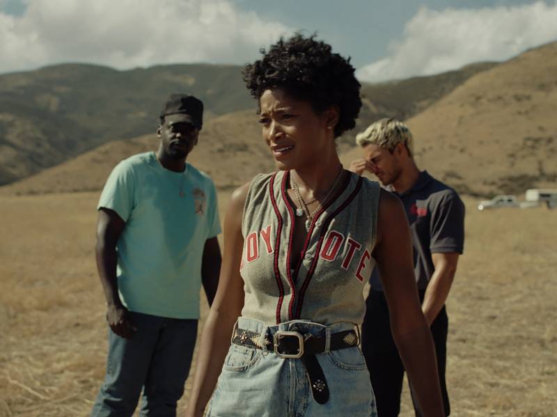 A slender Black woman with short hair wearing a sleeveless baseball shirt and high waisted blue jeans stands looking incredulous in the dessert. Behind her stand two men looking vaguely perplexed.