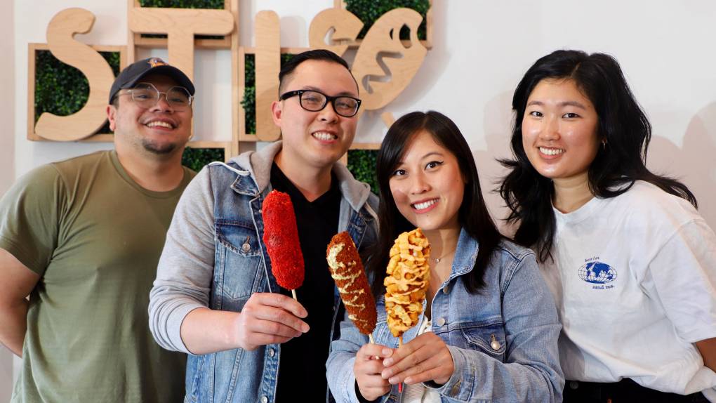 the owners of STIX hold two Korean corn dogs in their store, with two of their employees standing by their sides; behind them, STIX is printed on the wall