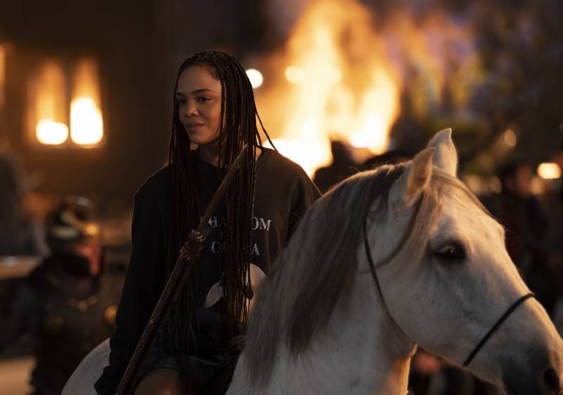 A Black woman with very long braids sits atop a white horse wearing a 'Phantom of the Opera' sweater. Flames are visible over her shoulder.