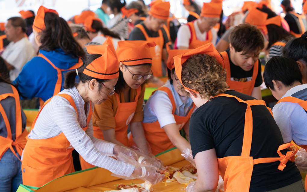 A crowded room of people in orange aprons and chef's hats, all preparing kimchi on the table in front of them.