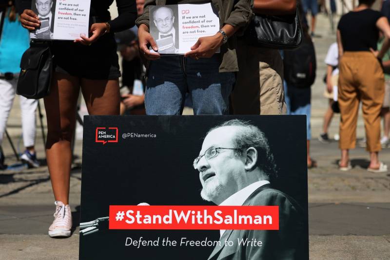A group of people stand holding signs that show Salman Rushdie alongside a quote from him: "If we are not confident of our freedom, then we are not free." In front of them is a large sign depicting Rushdie speaking. A red caption on the photo reads #StandWithSalman.