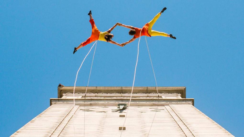 two dancers in red and yellow perform aerial arts off the side of a building against a blue sky