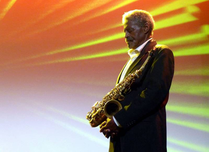 A man in a suit holds an alto saxophone against yellow and orange lighting