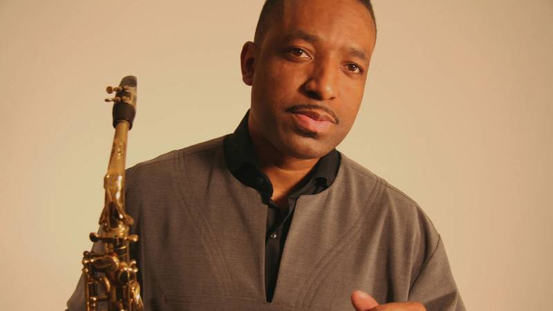 A man in a beige top holds an alto saxophone