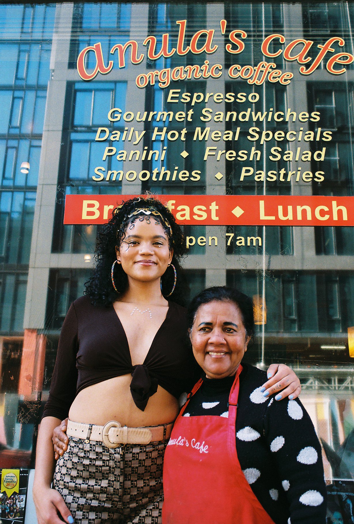 Anula Edirisinghe and her daughter Maya Rapier pose in front of Anula's Cafe; the sign in the window advertises the restaurant's offerings: "espresso, gourmet sandwiches, daily hot meal specials," and so on.