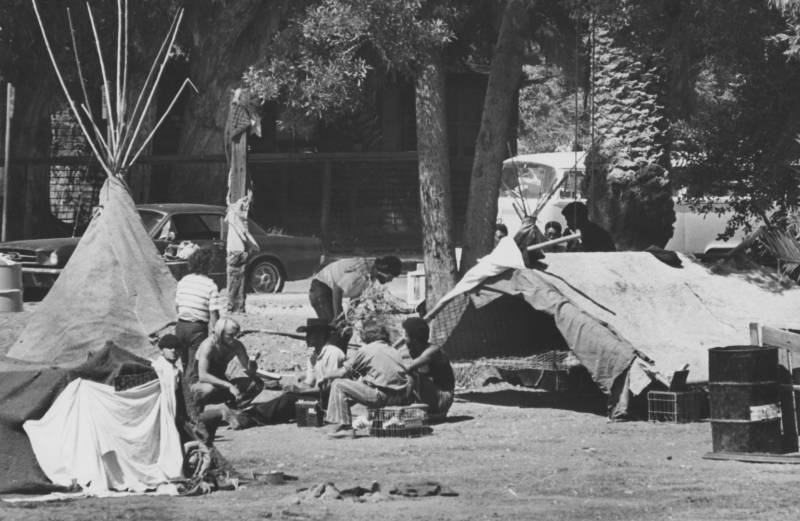 A group of men sit on crates and makeshift mattresses, next to two ramshackle tents in a park.