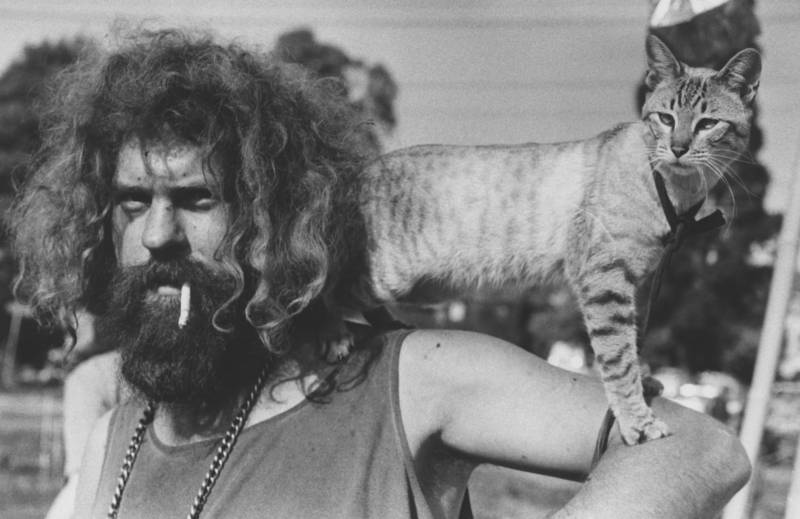 An unkempt man with tangled chin-length hair and a bushy beard smokes a cigarette while a grey striped cat stands on his shoulders.