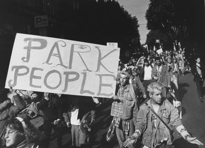 A large group of protesters march along a tree-lined street. Several hold up a large sign that reads 'Park People.'