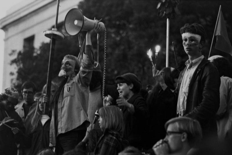 A masked demonstrator holds a megaphone aloft, as a speaker addresses a crowd.