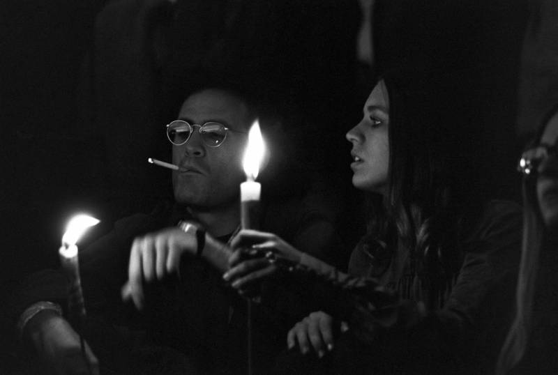 A young man wearing round spectacles smokes a cigarette and holds a lit candle. A young woman who long brown hair, also holding a candle, sits beside him.