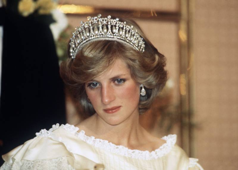 Princess Diana wearing a cream satin dress by Gina Fratini with an elaborate tiara and diamond earrings. Her head is tilted downwards, her face wearing a defeated expression.