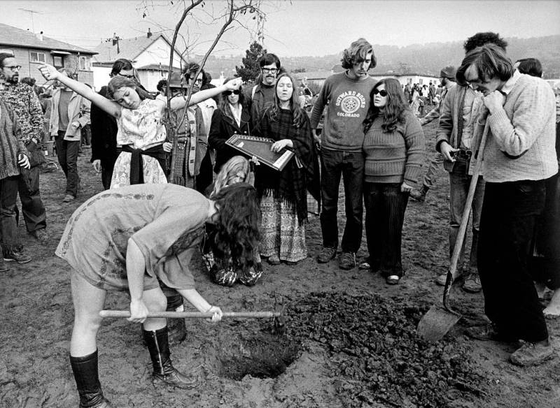 A woman bends down to dig a hole to plant a tree, while a gathered group watches on. One woman stands with her arms outstretched in joy. Another plays a zither.