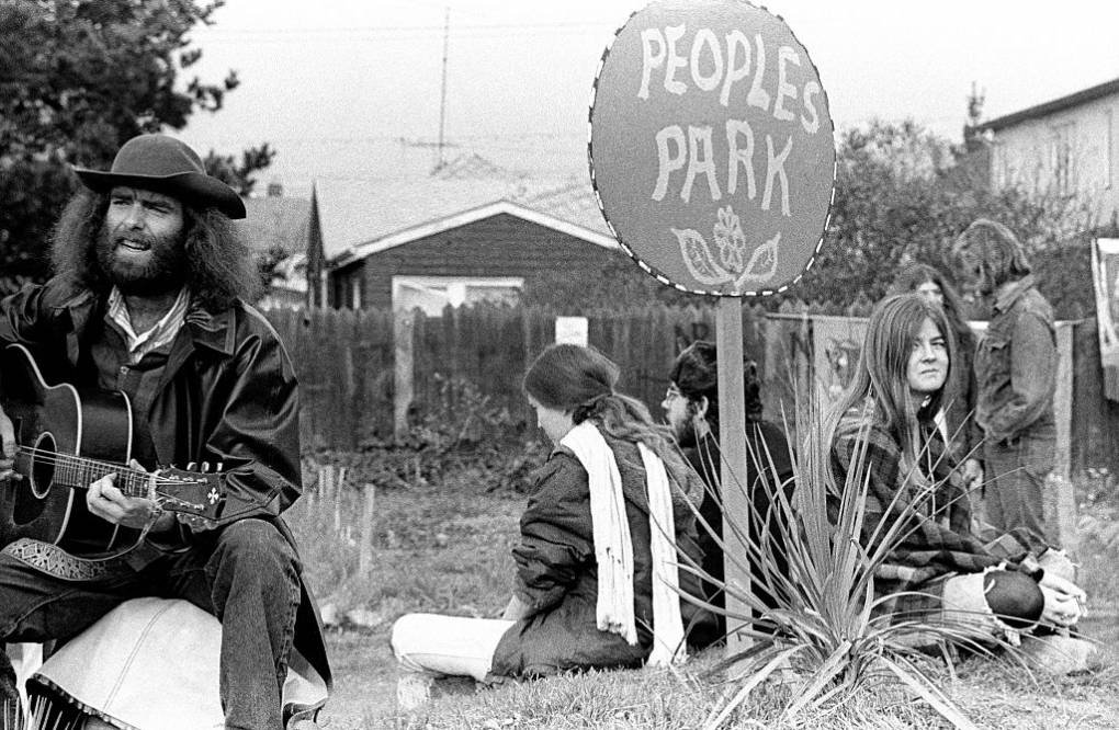 A man plays a guitar as others surround a sign that says People's Park. Two young women and a man sit nearby.