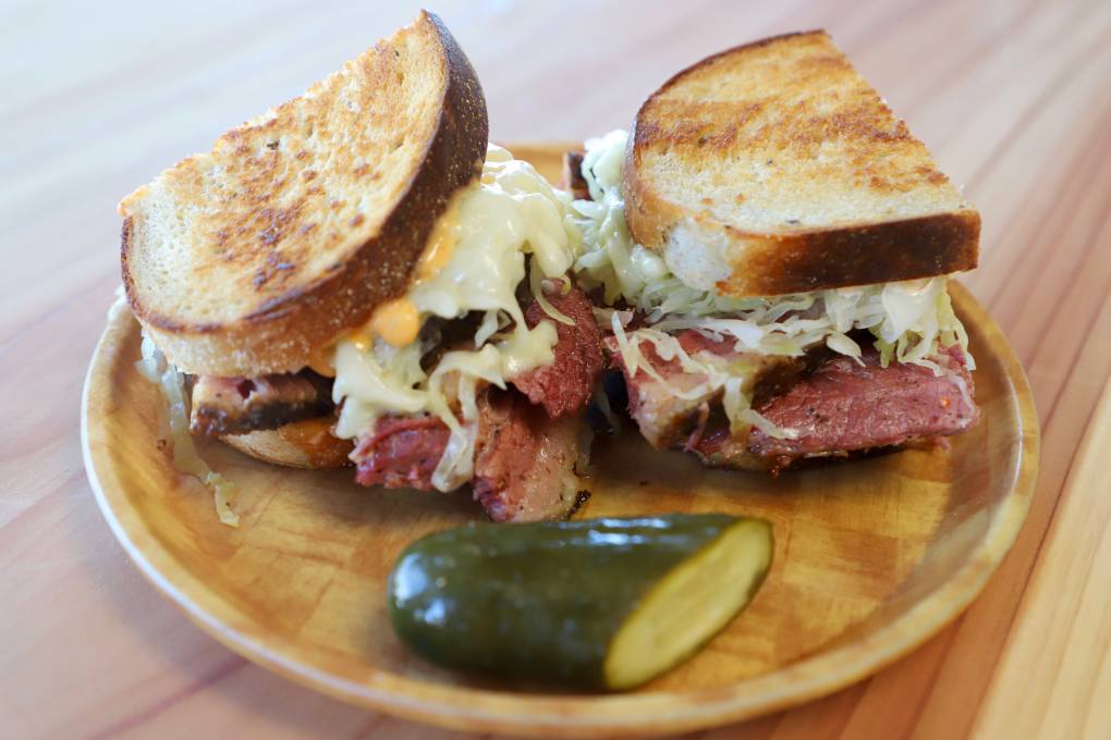a pastrami sandwich with coleslaw, cheese, and dressing served on a wooden plate with a pickle