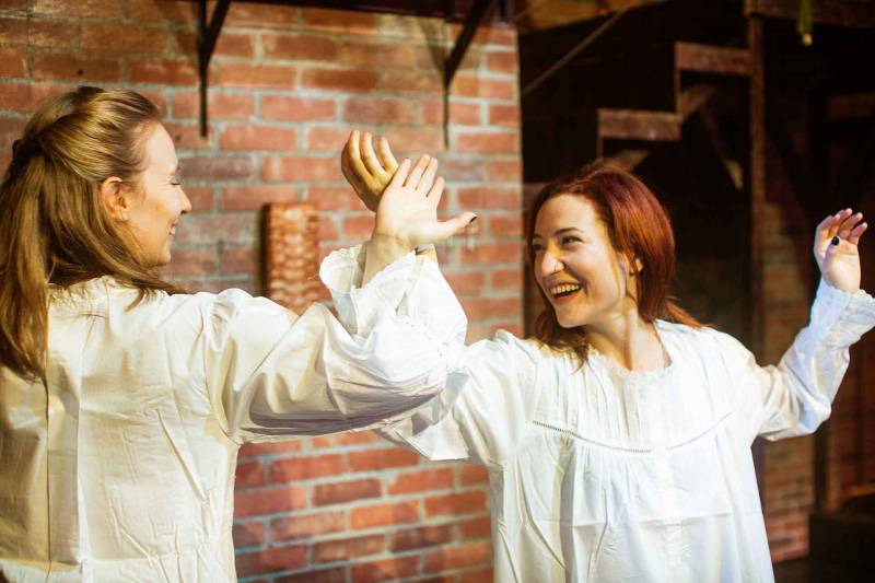 two women in white outfits dance in front of a brick wall in a play