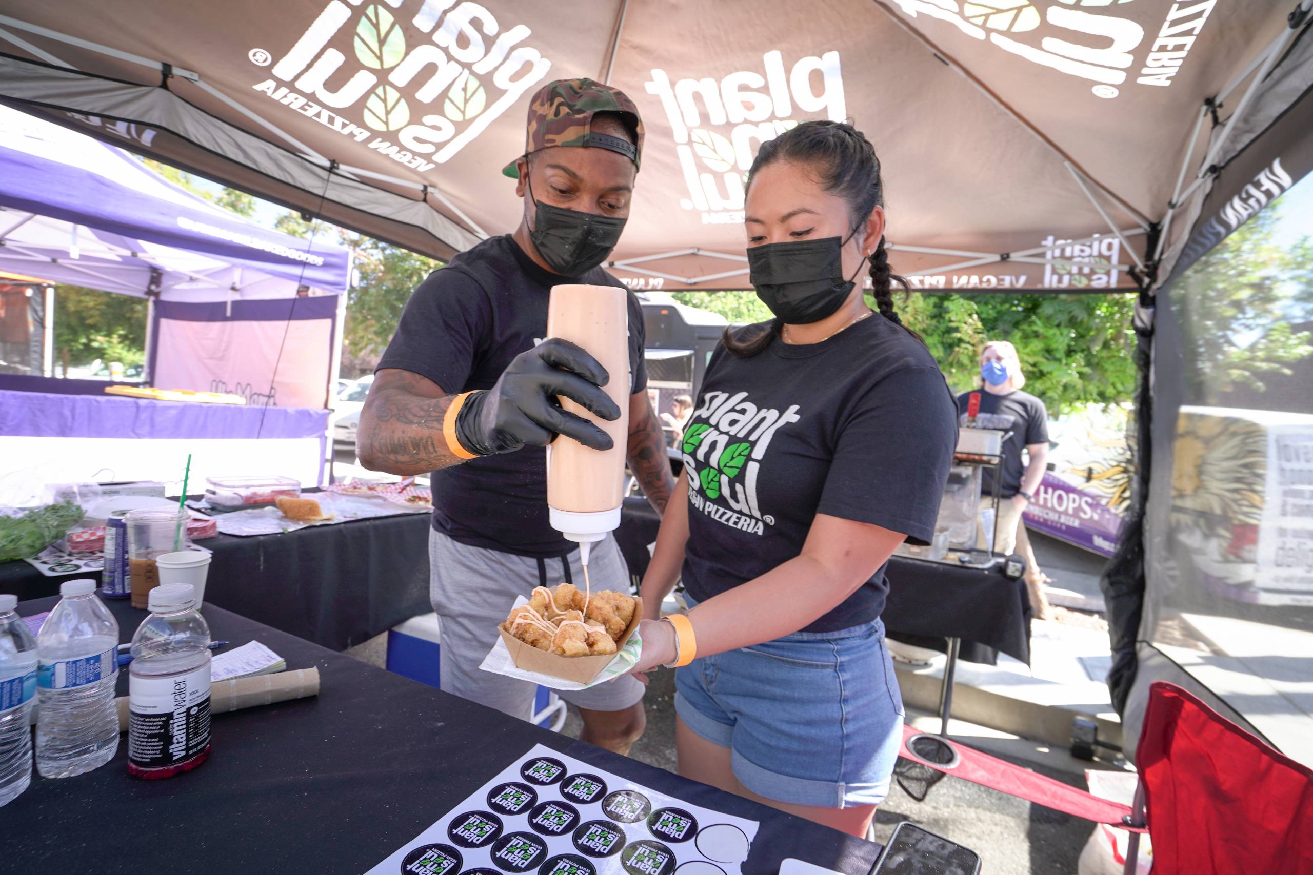 A man and woman in face masks squirt sauce on a paper tray of fried cauliflower. Their shirts read "Plant N Soul."