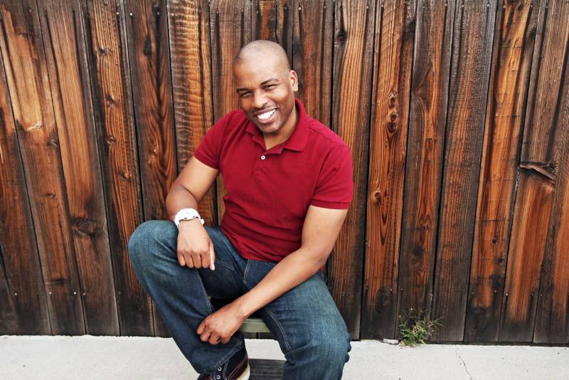 an African American man in jeans a red shirt smiles while posing in front of a wooden fence
