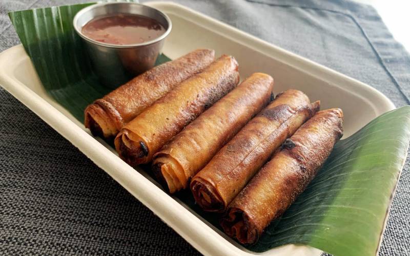 A cardboard try with five golden-brown lumpia, with dipping sauce on the side.