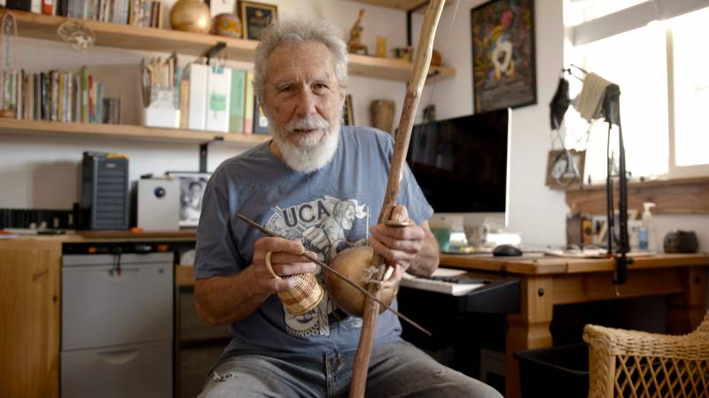 An older man with a white beard and mustache is looking into the camera while playing the berimbau.