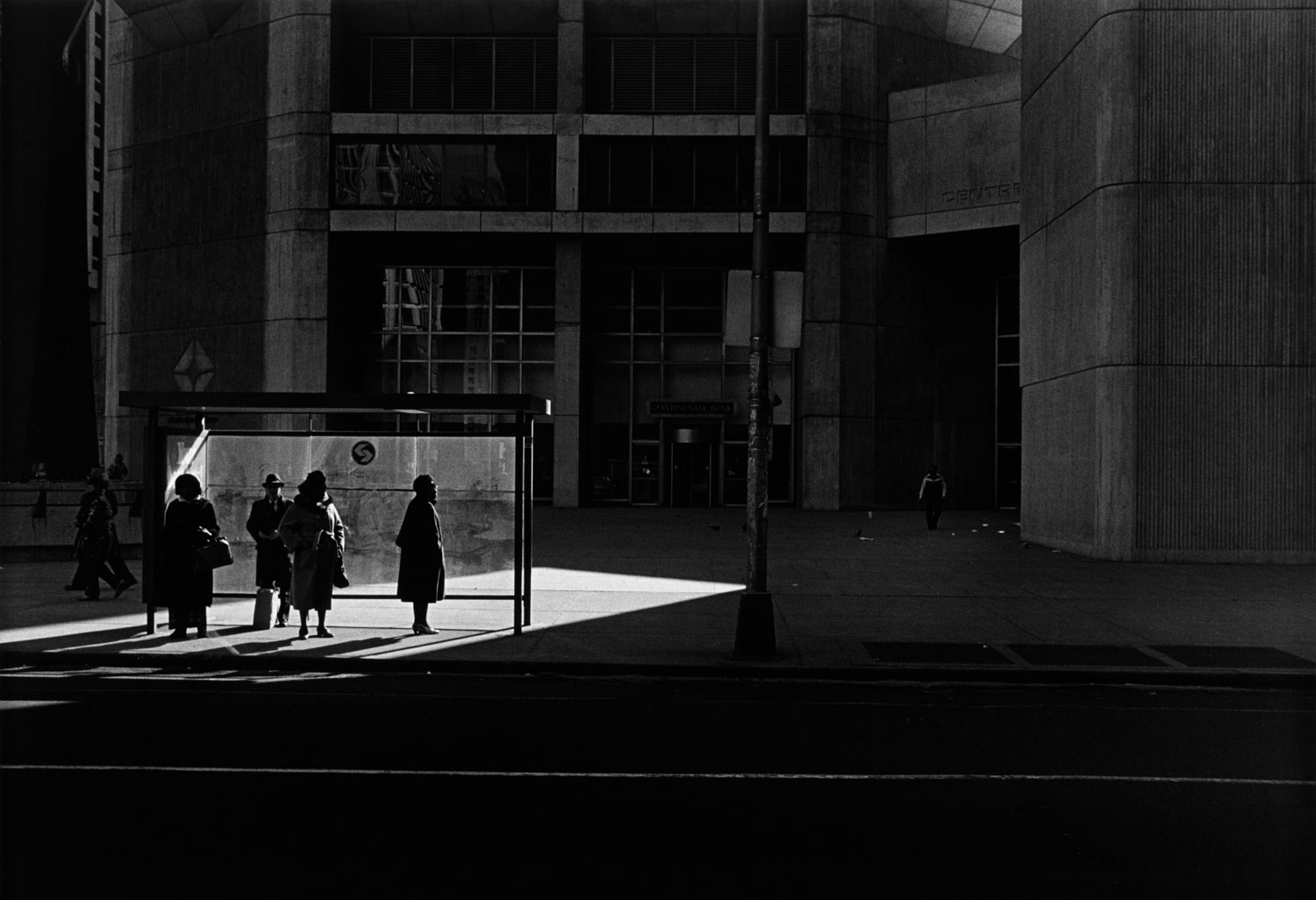 Black-and-white photo of people waiting for bus in angled patch of light against dark city landscape