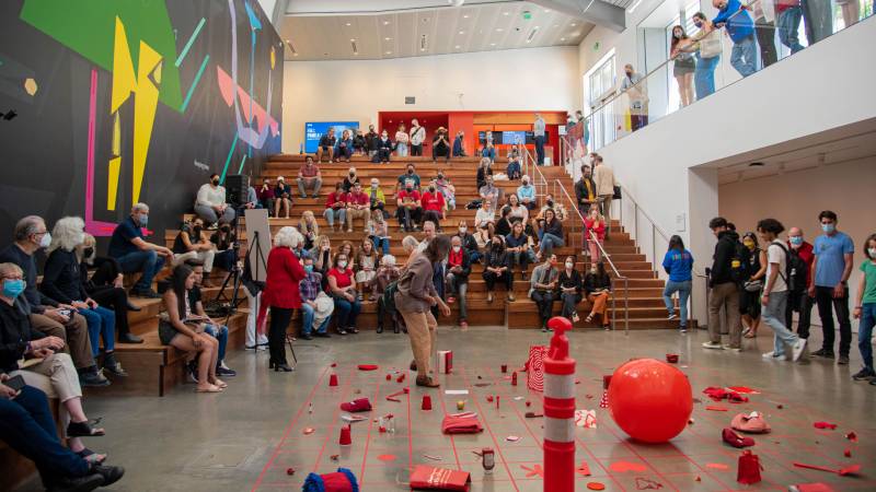 Grid arrangement of red objects on floor in front of stadium seating filled with people in museum atrium.