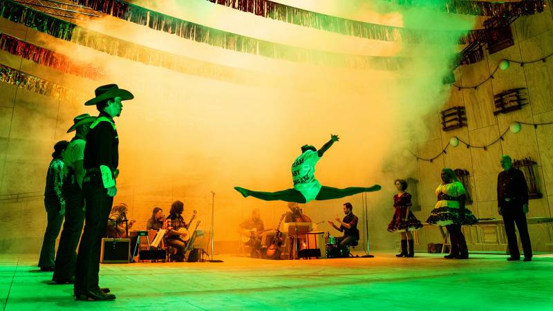 A young black female dancer leaps in the air as yellow smoke pours out over the play's cast