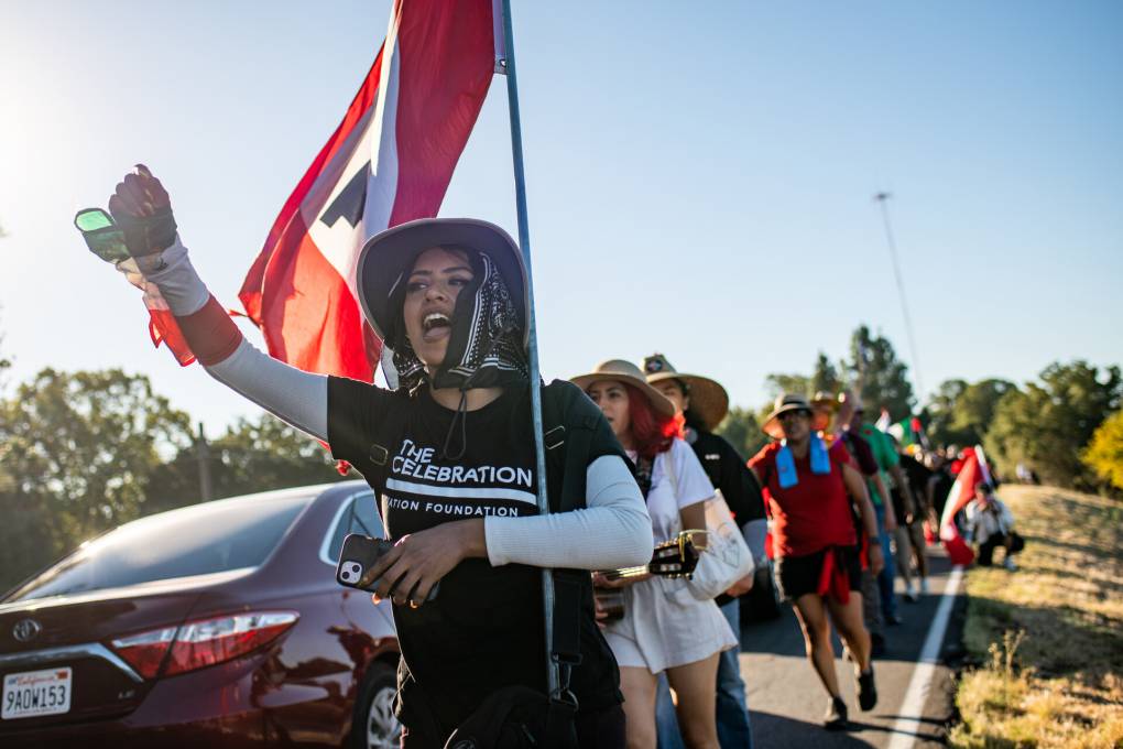 An activist holding an red-and-white United Farm Workers flag raises her fist as she leads a group of marchers walking along the highway.