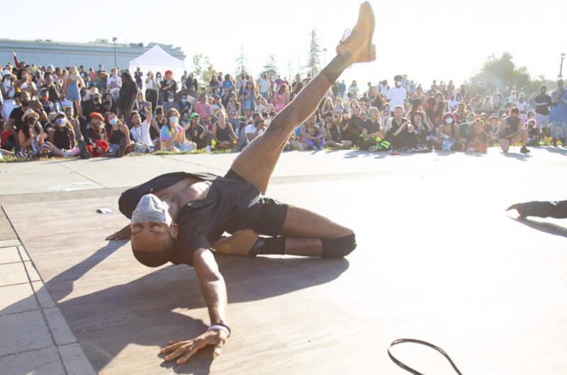 A Black dancer in open black shirt and tight black shorts, lays on a wooden platform, arms outstretched dramatically, with one leg kicked upwards and pointed out. Crowds watch on from the sidelines.