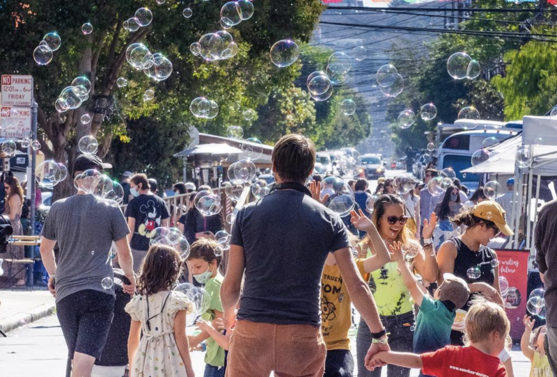 A father and son walk hand-in-hand towards a group of parents and children playing with bubbles. Behind them, various stalls can be seen lining the street.