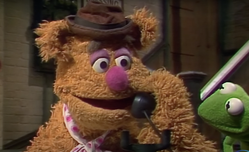 A muppet bear wearing a white and pink dotted silk neck scarf and small brown hat talks on the telephone.