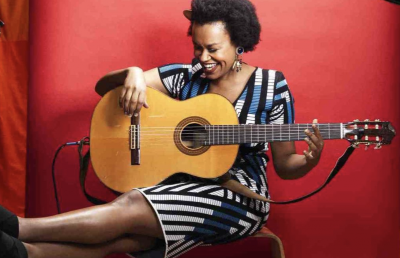 A Black woman in black and white striped dress, sits on a stool with her feet up, laughing as she holds an acoustic guitar on her lap.