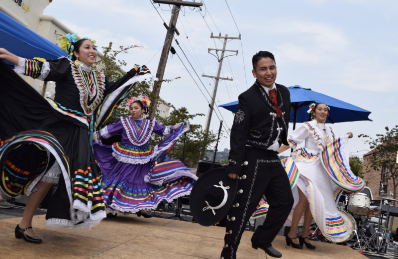 Dancers - three female, one male - wearing traditional Mexican clothes smile as they perform on a small stage.