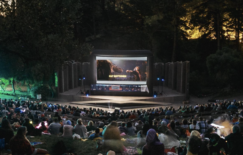Many people gather on the slanted lawn in front of a large screen surrounded on all sides with trees.