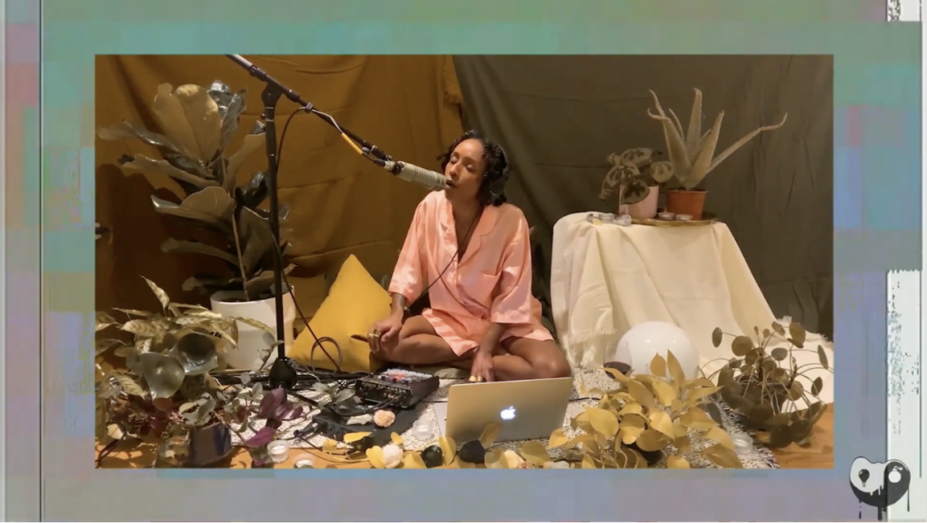 a woman in a pink robe sits on the floor surrounded by plants, singing into a microphone